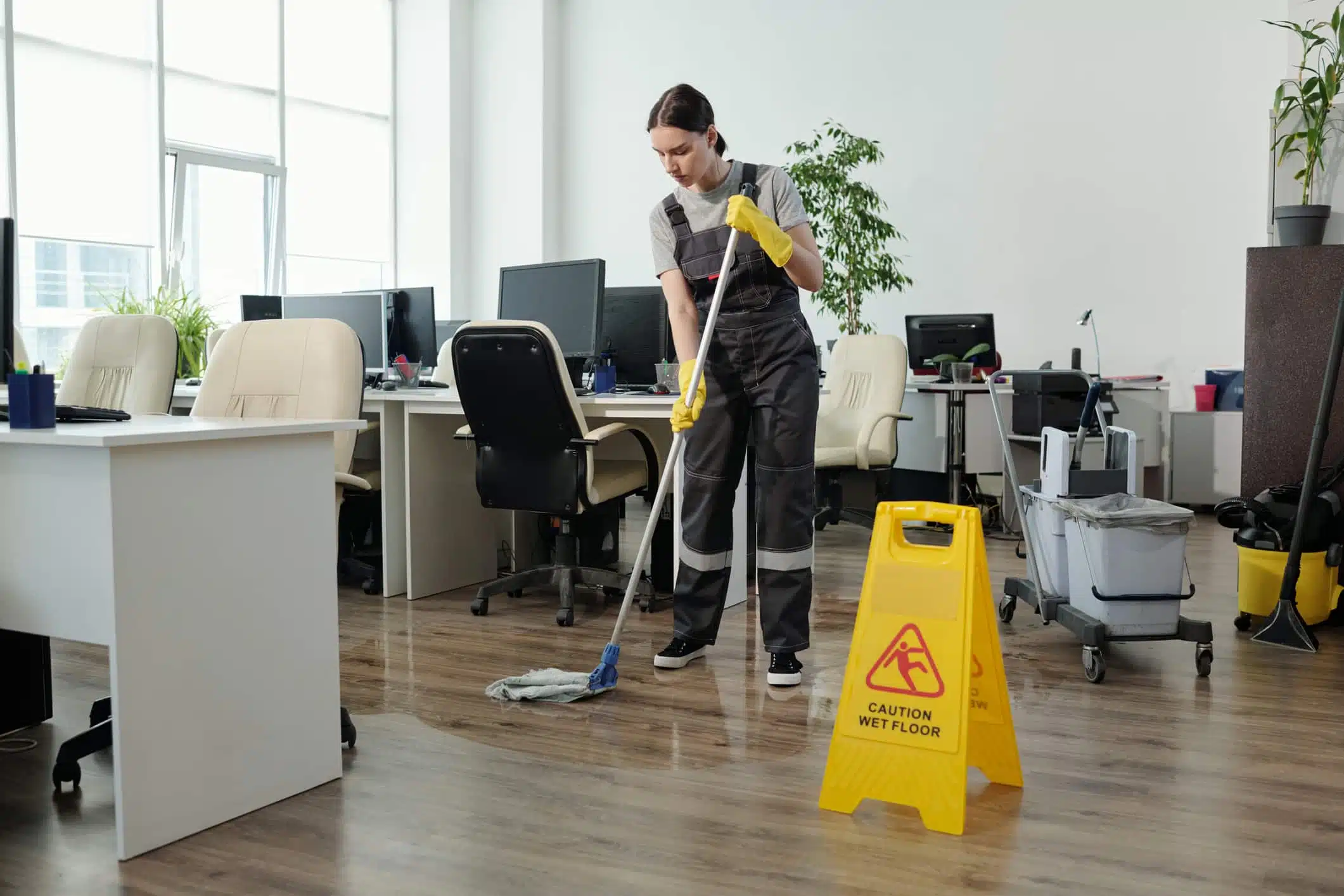 Young female cleaner in workwear using mop while cleaning floor in large modern openspace office with yellow plastic signboard in front. Article refers to non-slip coatings in teh workplace